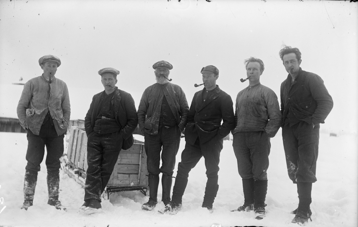 Seks menn på jakt, alle med piper og med fotografen Kjell O. Moe til høyre. Ei slede i bakgrunnen.