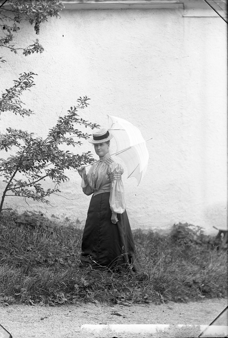 Porträtt av Jenny Rosenborg. Hon står med ett parasoll mot en husfasad, möjligen vid Bergsgatan 58. Hon bär en stråhatt, ljus blus och mörk fotlång kjol.