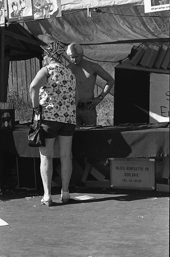 Marknad på okänd plats. En kvinna i kortbyxor, ärmlös topp och hatt står vid ett marknadsbord. Knallen har bar överkropp.