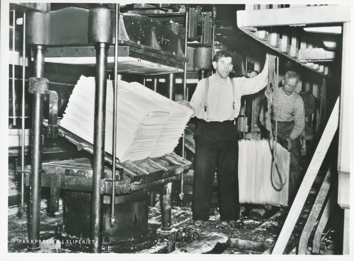 To menn iført skjorte og bukser med seler arbeider med pakkpresser som presser tremasseballer.