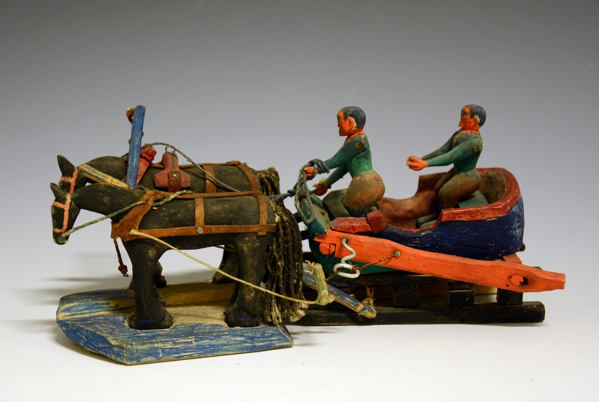 Prot: Leke. Utskåret i tre. Slede med to personer oppi med to hester forspent. Malt i brun, grønn og rød farge.