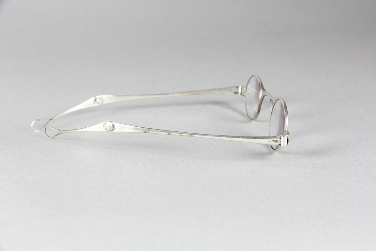 Glasögon, mindre glas med smala bågar av silver. Två ledade skalmar som utåt avslutas med droppformade öglor.