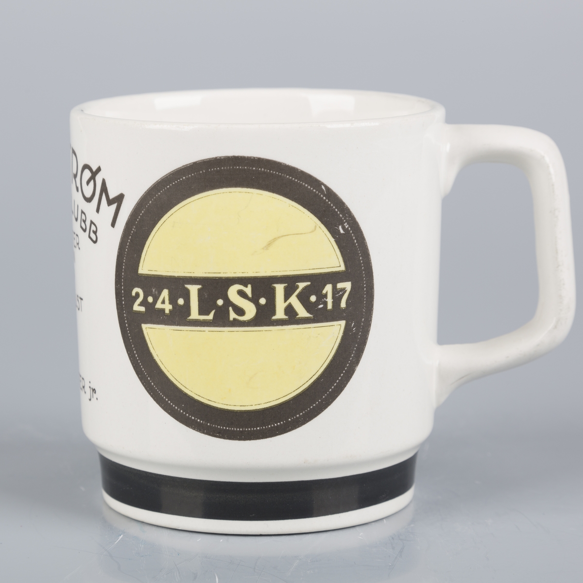 Krus med hank med LSK-logo og tegning av en pokaler. Stempel under inkluderer en flinthammer.