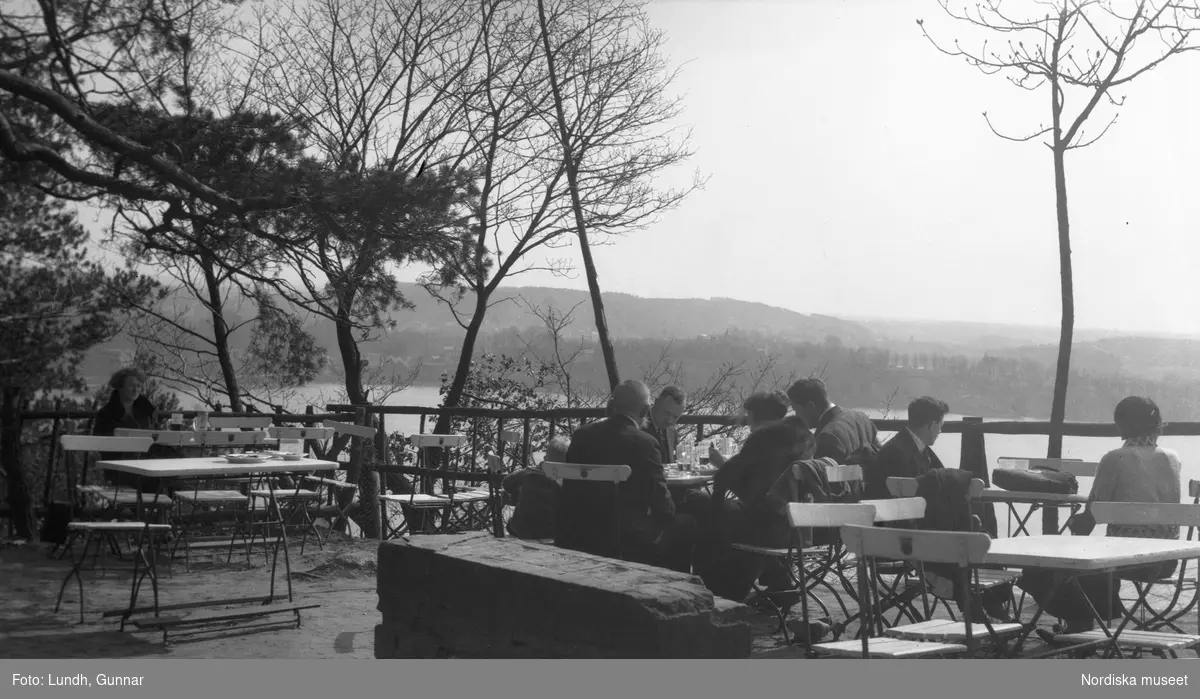 Motiv: Utlandet, Märkische Schweiz 102 - 108 ;
Kvinnor och män sitter på en uteservering vid en sjö, landskapsvy med träd - vass och sjö.