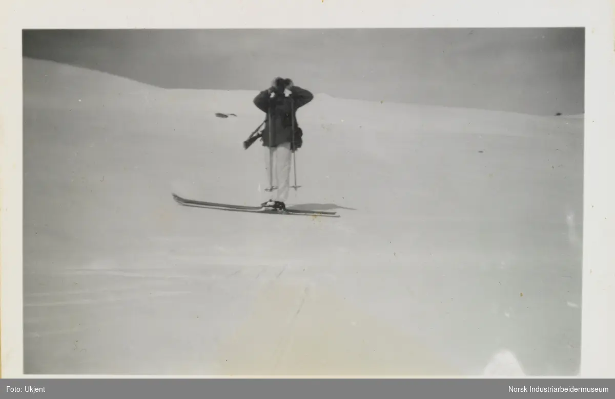 Mann med gevær på rygge står på ski i fjellet og ser med kikkert