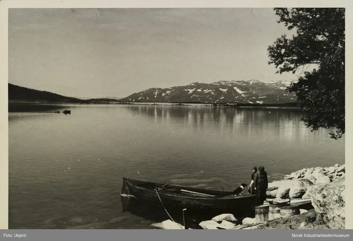 Terjei og Oline Skinnarland står ved snekke og pråm i vannkante på Møsvatn ved Skinnarland. I båten sitter en hund. Ute på innsjøen sees en annen båt