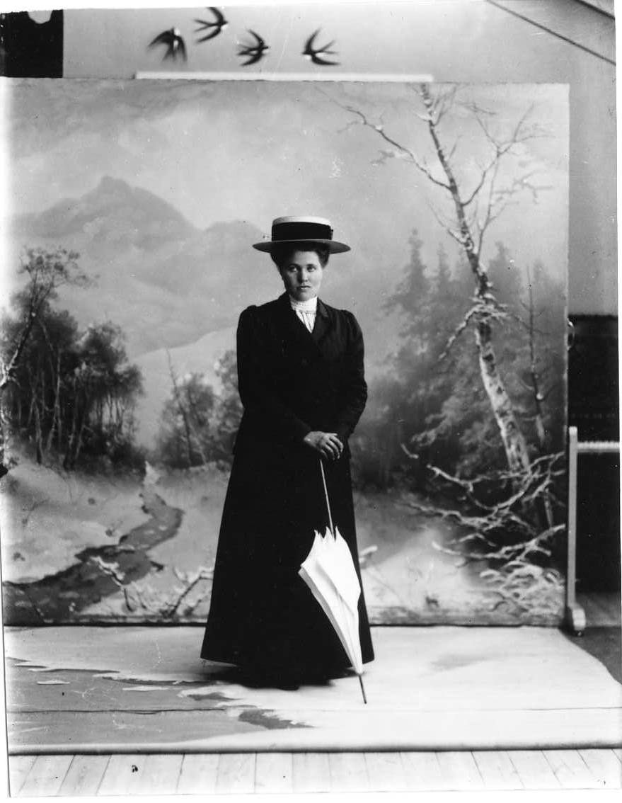 Porträtt av Anna Johansson från Fallet i Gränna socken. Hon står med ytterkläder, hatt och parasoll framför en vinterfond.
