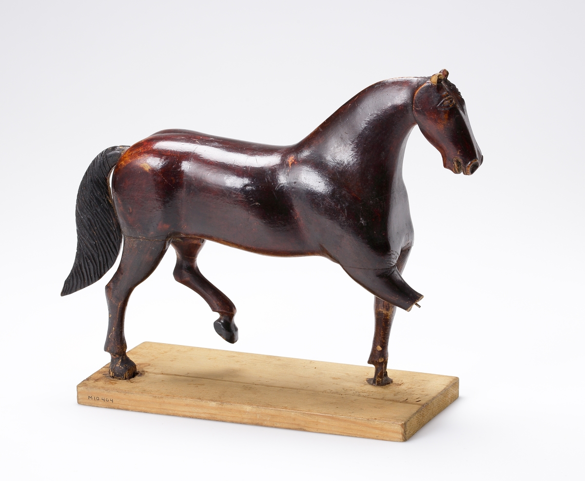 Leksakshäst av brunt trä med remtyg och tömmar av ljusbrunt läder (saknas). Står på två ben på en omålad träplatta.

Inskrivet i huvudkatalog 1939.