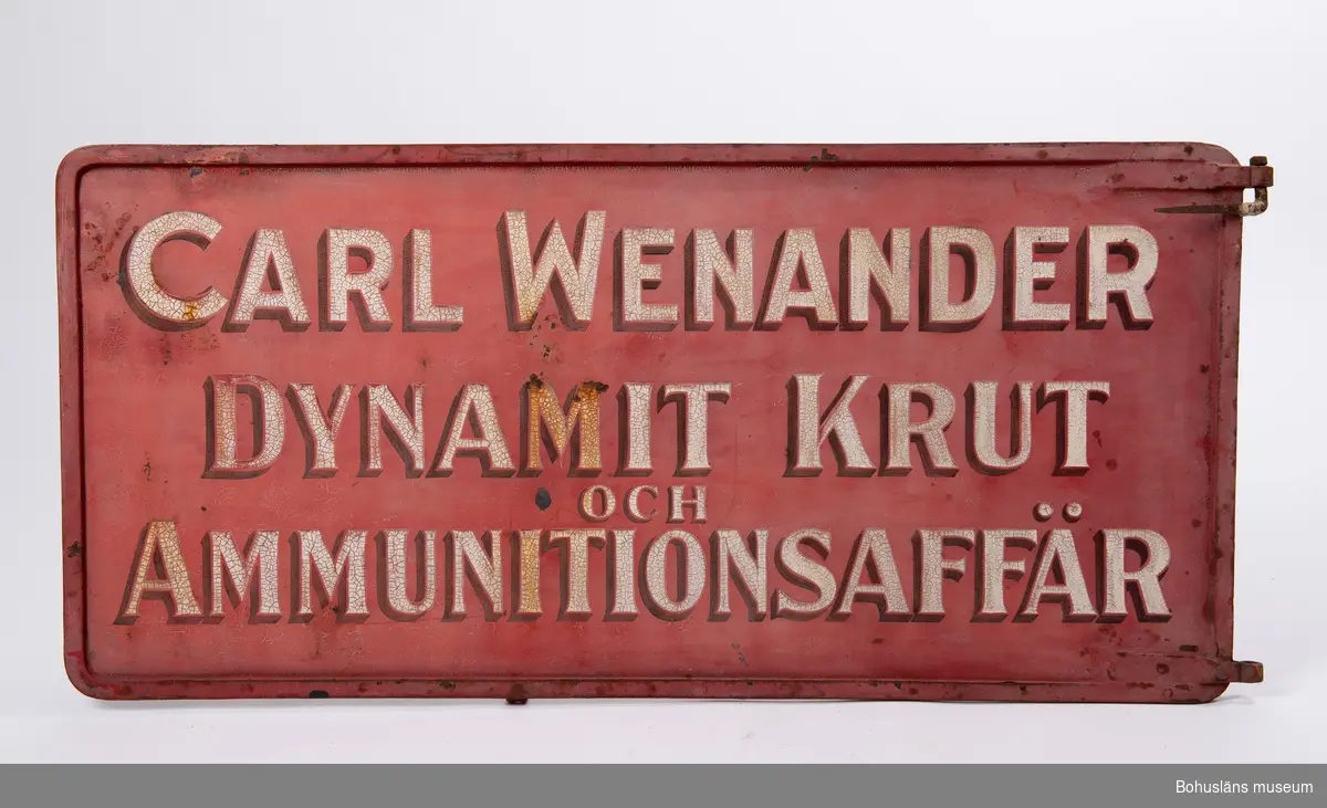 Text å båda sidor i vitt och svart mot botten i engelskt rött: 
"Carl Wenander. Dynamit, -Krut och Ammunitionsaffär."
