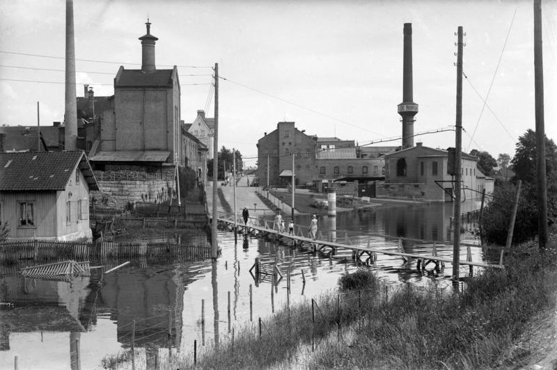 Svart-hvitt fotografi som viser en vannfylt gate med provisorisk bro over, et lite plankehus til venstre og to store murbygninger; Hamar bryggeri og melkefabrikken med en høy, rund mursteinspipe.