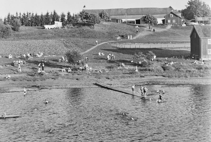 Svart-hvitt fotografi som viser mange mennesker som bader og soler seg. Det er gress på bakken og en lang brygge utover i vannet. I bakgrunnen vises Storhamarlåven.