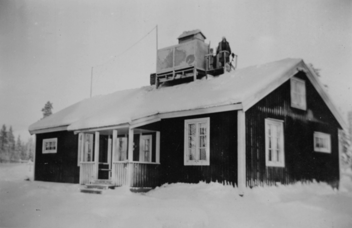 Exteriör av militärförläggning för 91:a ls-kompaniet för luftbevakning i Tellejåkk, Kåbdalis under beredskapen, 1942. Ett hus med luftbevakningstorn på taket. På taket står en kvinnlig luftbevakare.