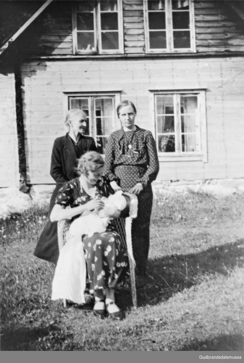 Tre kvinner samlet på tunet på Nørdre Plassen, den ene kvinnen sitter i en stol og holder dåpsbarn. Einar Sandgro i dåpskjole.
