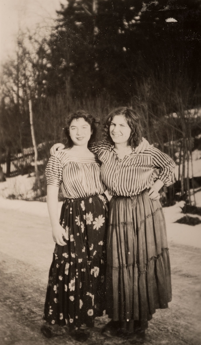 Två romska kvinnor står på en landsväg i Storvik vintern 1947. De har utslaget hår och är klädda i kjol och blus. I bakgrunden syns ett staket mot ett skogsparti.