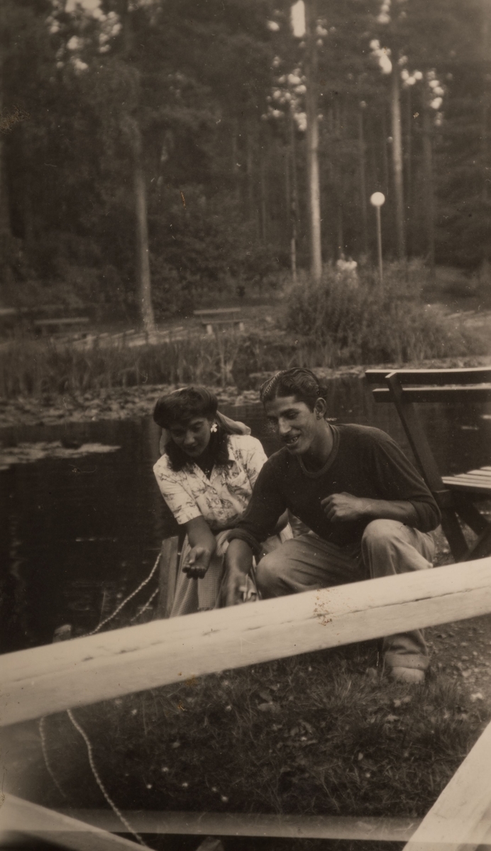En romsk kvinna och hennes man sitter vid vattnet i Stadsparken, Falun augusti 1950. På andra sidan vattnet syns parkbänkar, träd och parkbesökare.