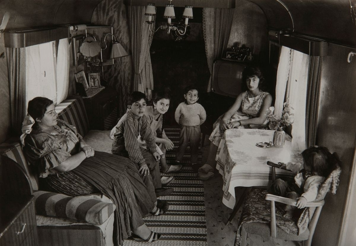 Två kvinnor och en samling barn sitter i en bostadsvagn. Bilden är tagen i Skarpnäck, troligen  i något av de läger som irodningsställdes av Stockholms stad i slutet av 1950-talet.