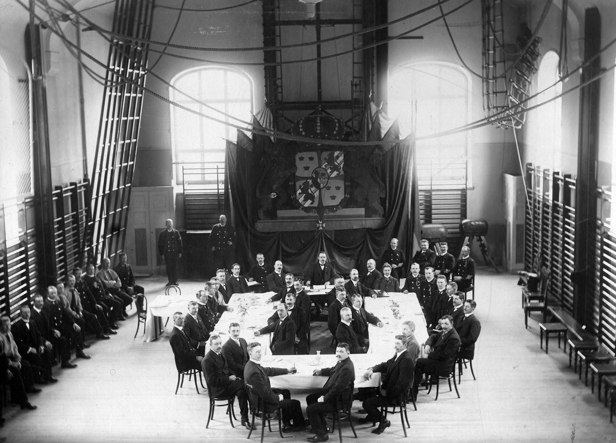 Brandkårsförbundets årsmöte i Stockholm omkring 1900. Ett antal män sitter vid ett dukat bord och längs väggarna i en gymnastiksal.