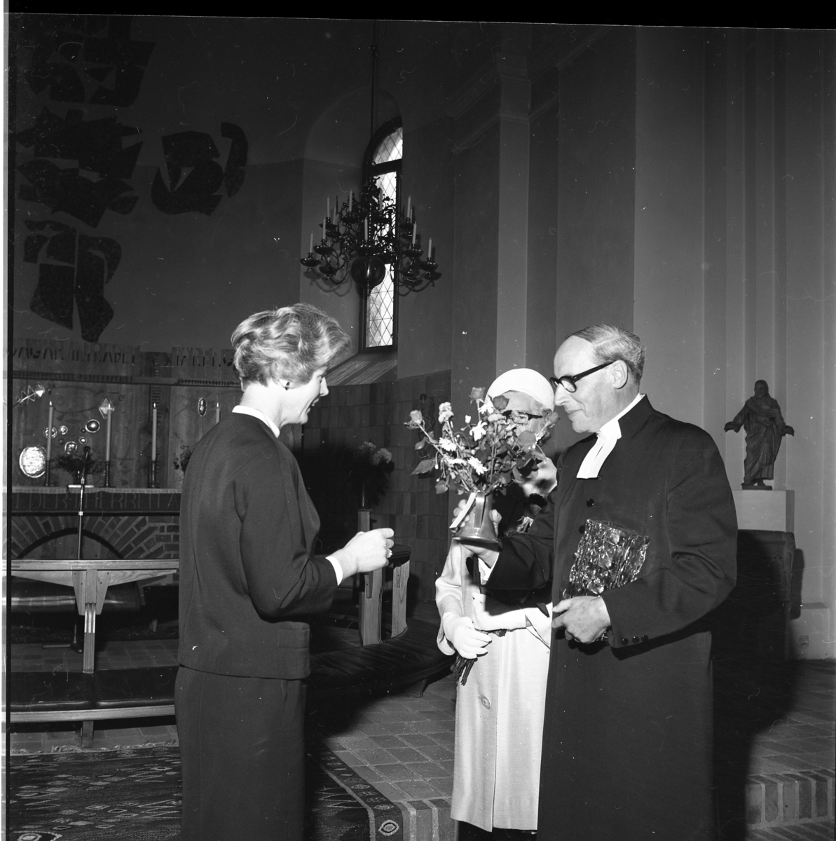 En prost glasvas och blomsterbukett i händerna samtalar med en kvinna. En annan kvinna står strax intill prosten. De befinner sig i Lommaryds kyrka.