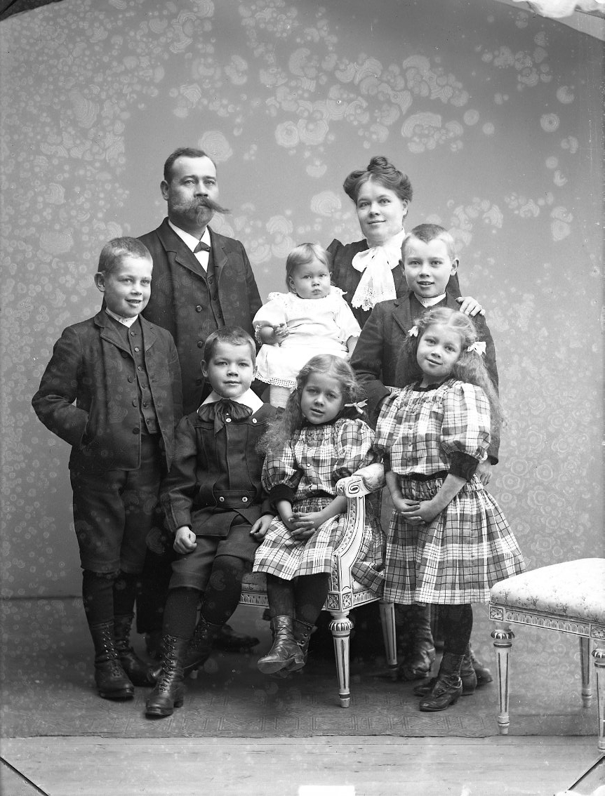 Gruppbild med familjen Johansson. Sex barn - tre flickor och tre pojkar - står och sitter framför herr och fru Johansson. De äldre flickorna har storrutiga klänningar och rosetter i håret. De ler.