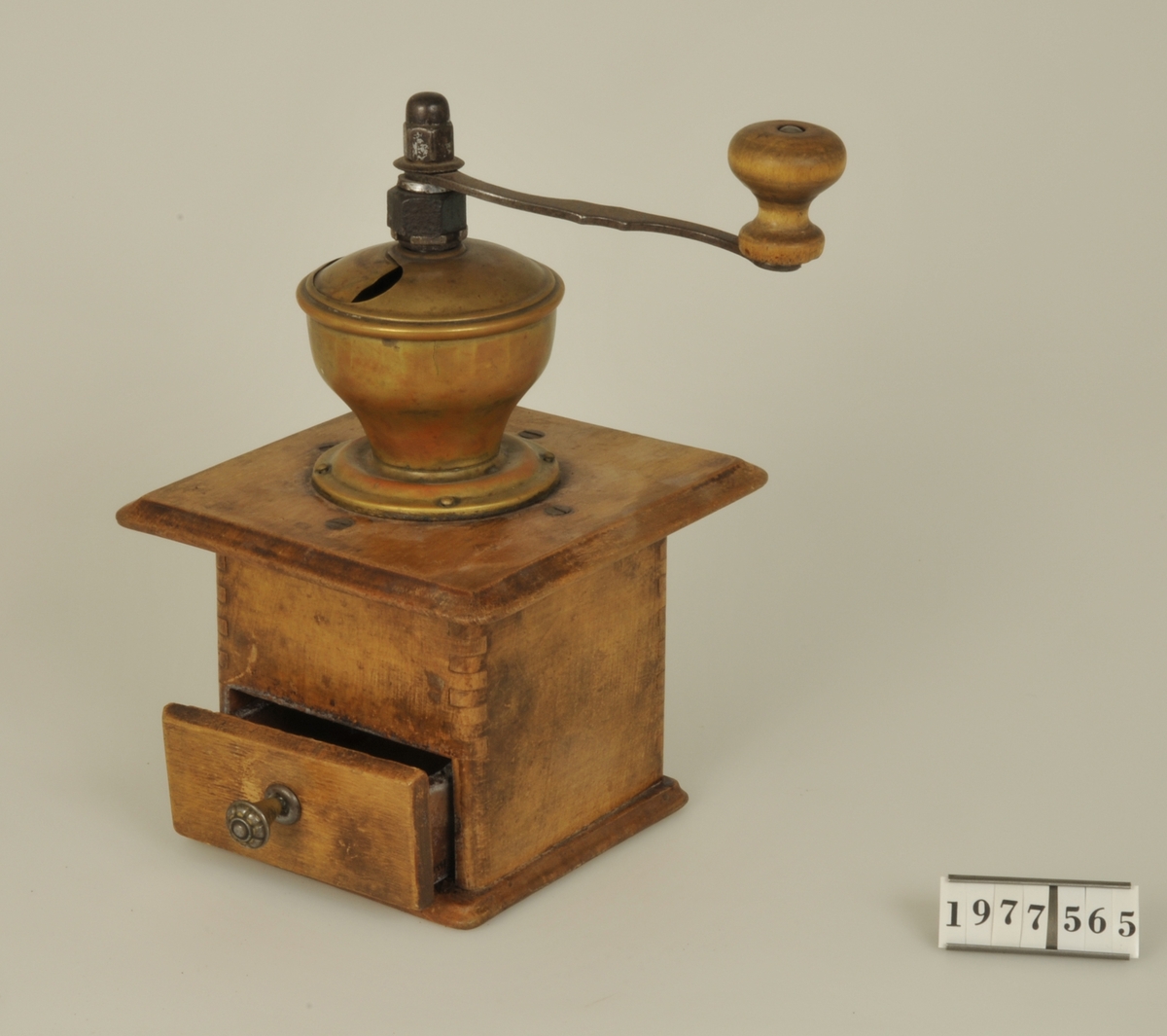 
Kaffekvarn med fyrkantig låda av trä, skål av mässing samt vev av järn med handtag av trä.