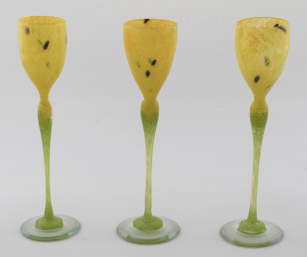 Tre høye drikkeglass med tulipanlignede utforming. Eggeformet kupa i gult halvgjennomskinnelig glass med sorte prikker. Stetten er utført i lysegrønt opakt glass, som hviler på en transparent fot.