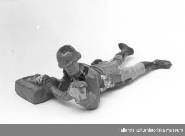 Leksakssoldat av trämassa på ståltrådsskelett i tysk uniform. Soldaten ligger ner och matar ett band av kulor. Målad i grönt, svart, guld och skärt.