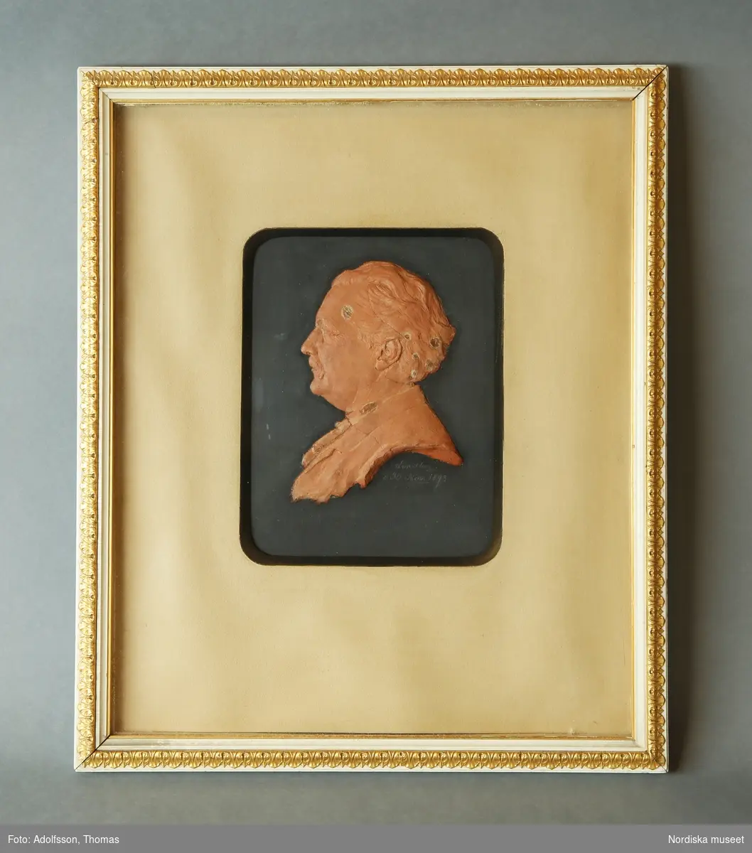 Inramat porträtt i profil av Artur Hazelius. Utförd i rödfärgad gips, infäst i mot bakgrund.