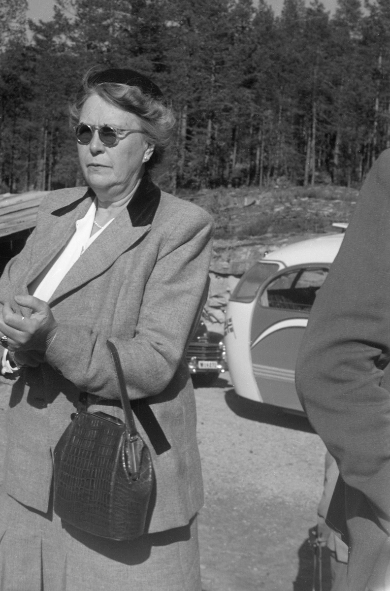 Ingeborg Bødtker (1895-1985), fotografert under Norsk fløtningsforbunds befaring i 1952. Hun var gift med [Jacob Gottfrid] Otto Bødtker (1888-1966), som først var fløtingsdirektør i Drammensvassdraget (1915-1925), deretter i Skiensvassdraget (1925-1928). Følgelig hadde hun antakelig en slags vertinnefunksjon da organisasjonen ektefellen var direktør for samlet norske fløtingsfunksjonærer og styremedlemmene i Nordisk fløtningsråd til konferanse med innlagt befaring og årsmøte høsten 1952.  Ingeborg Bødtker var kledd i ei drakt med skjørt og jakke som på fotografiet framstår som lysegrå, med unntak av den bakre delen av kragen, som er mørk. Under jakka hadde hun en kvit bluse. Ingeborg Bødtker hadde alpelue på hodet, solbriller på nesa og håndveske over venstre underarm. Ellers var hun pyntet med perlekjede og perleøredobber. I bakgrunnen ser vi bakparten på ekskursjonsbussen og fronten på en bil av merket Plymouth, antakelig en årsmodell fra perioden 1946-1948.