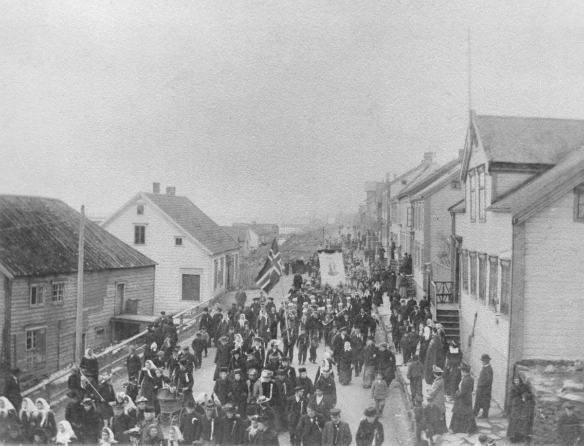 17. mai-tog i Vadsø i 1905 med flagg og musikkorps. Sivertsengården (Ekviperingen) til høyre.