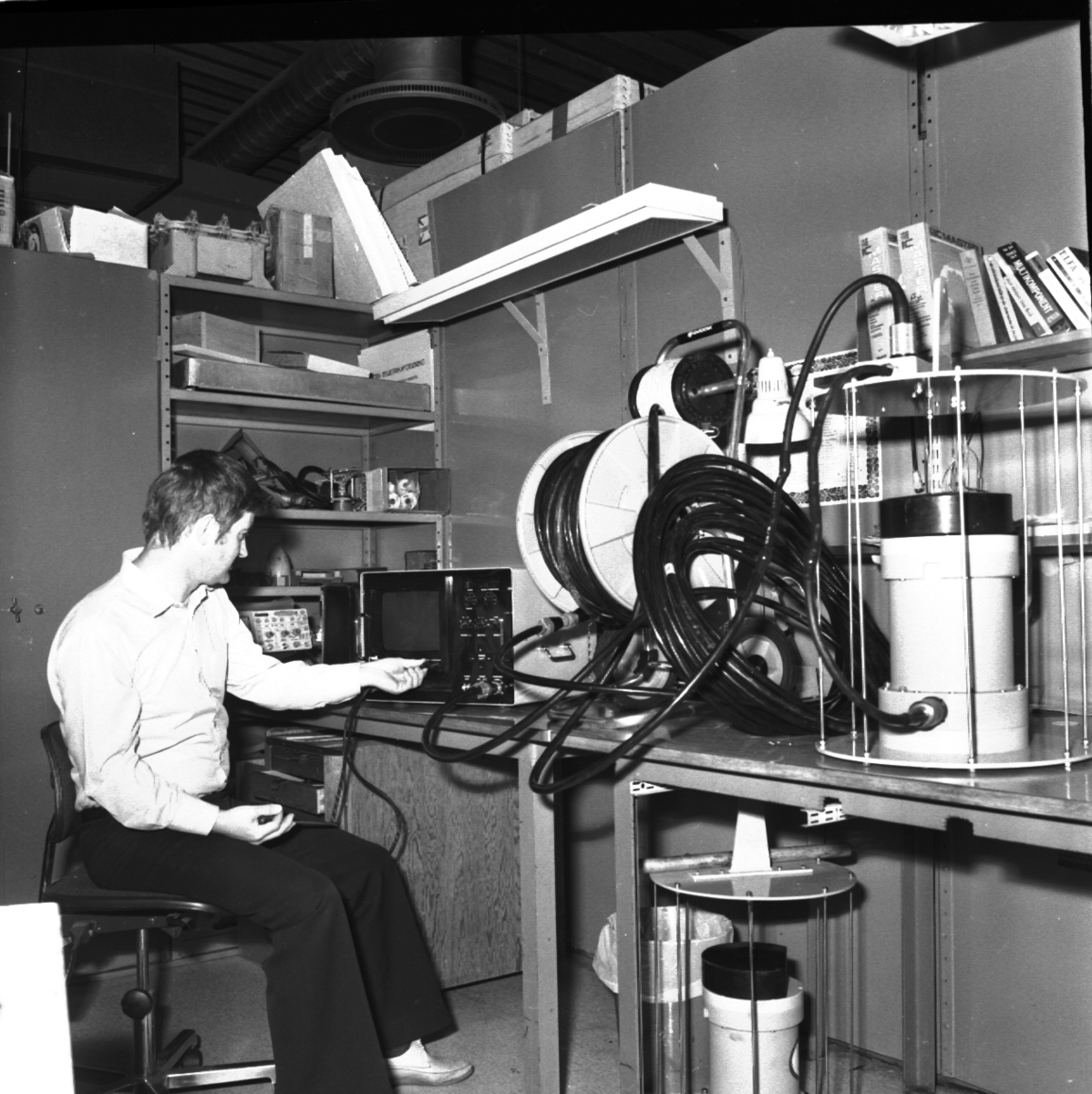 En man sitter på en snurrstol framför en maskin med skärm och kopplar in en sladd. Han sitter i ett rum med sladdar och maskiner på hyllor och bord.
