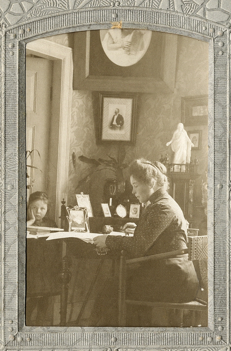 En kvinna i prickig blus och mörk kjol sitter och arbetar vid ett skrivbord. Till vänster syns en liten flicka. I hörnet ser man ett foto av en man (kvinnans make, trävaruhandl. Edvard Andersson (1875-1934).