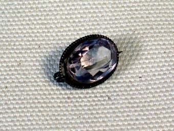 Oval genomskinlig slipad sten av något slag i ram av tunt flätat band av mässing. På baksidan en säkerhetsnål med vilken man fäster broschen på plagget.