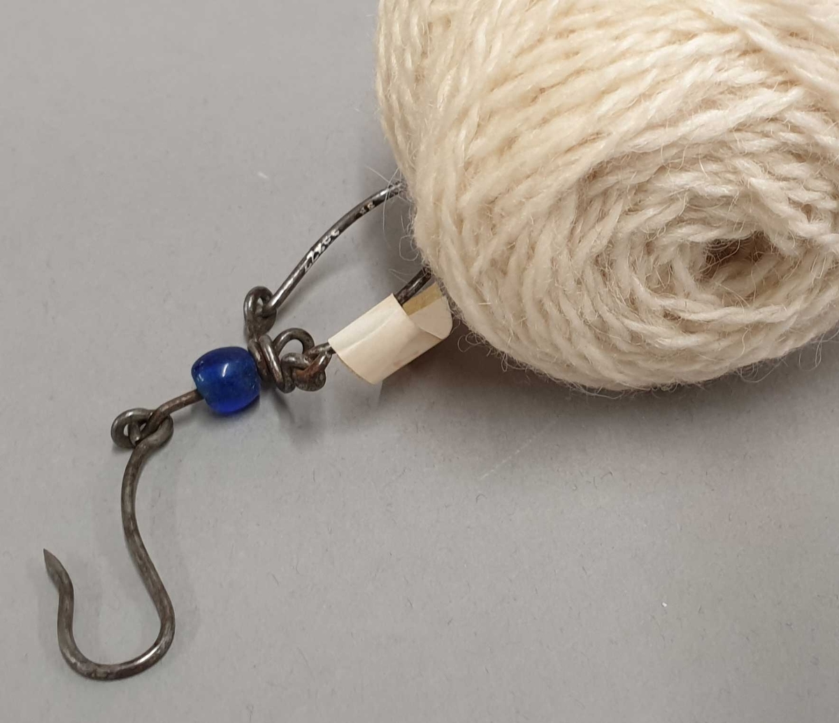 Nøstekrok som består av en krok og en bøyle som kan åpnes forbundet med et mellomstykke laget av tråd. På mellomtråden er tredd innpå en liten blå glassperle. Krok og bøyle kan dreie fritt rundt hverandre.