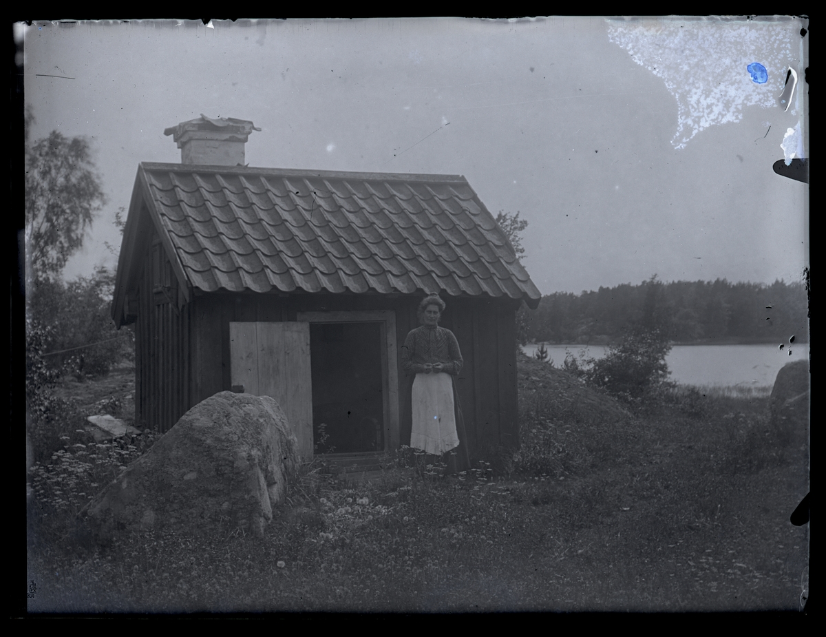 Byggnad, troligen tvättstuga, samt kvinna. Lillhärad socken, ca 1910.