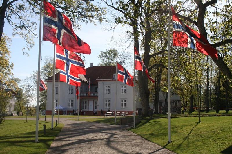 Et fotografi av Eidsvollsbygningen med en rekke av norske flagg på flaggstenger på begge sider av grusgangen opp mot huset. Bildet er fra 17.mai