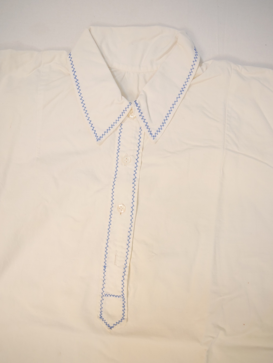Form:  Sid kjole med rette sider, isydde lange ermer. Skjortekrage, ermkragar og stolpe kanta med blå siksaksting. Ein perlemorsknapp på kvar av ermkragane, ein av fire perlemorsknappar på stolpen manglar.
