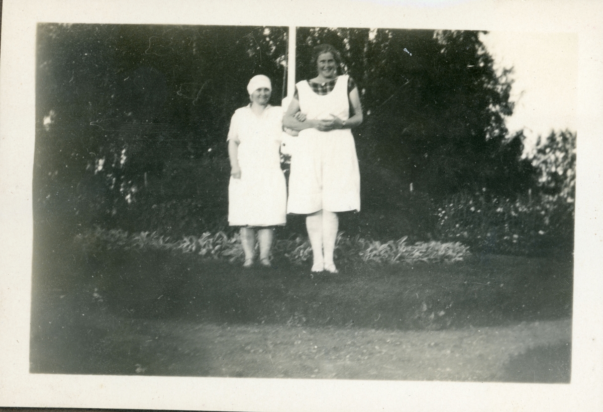 Elever fra husmorskolen på Skansgården, Kongsvinger, 1928. To unge kvinner i kjøkkenklær/arbeidsklær foran flaggstang og blomsterbed.