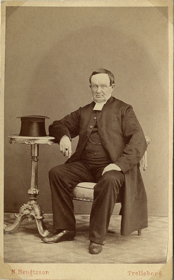 Porträttfoto av en man i prästdräkt m.m. Han sitter på en stol och vilar ena armen på ett pelarbord, där det ligger en hög hatt. 
Helfigur. Ateljéfoto.
