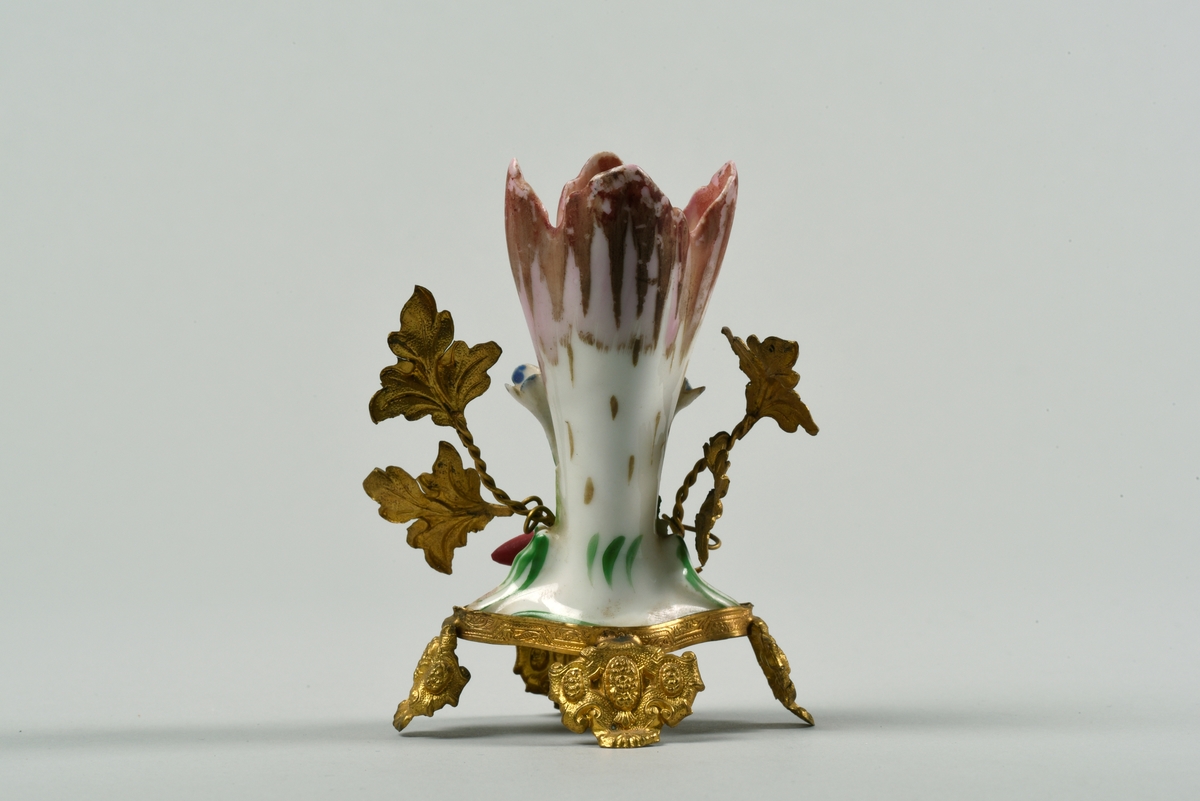 Porslinsvas med ställning i guldmålad metall. Vasen är formad som en blomsterkalk med dekor av platsiskt gjorda blommor och blad i rokokostil. Vasen står i en ställning i form av blad med tvinnade stjälkar. De fyra fötterna är i utformade som sköldar.