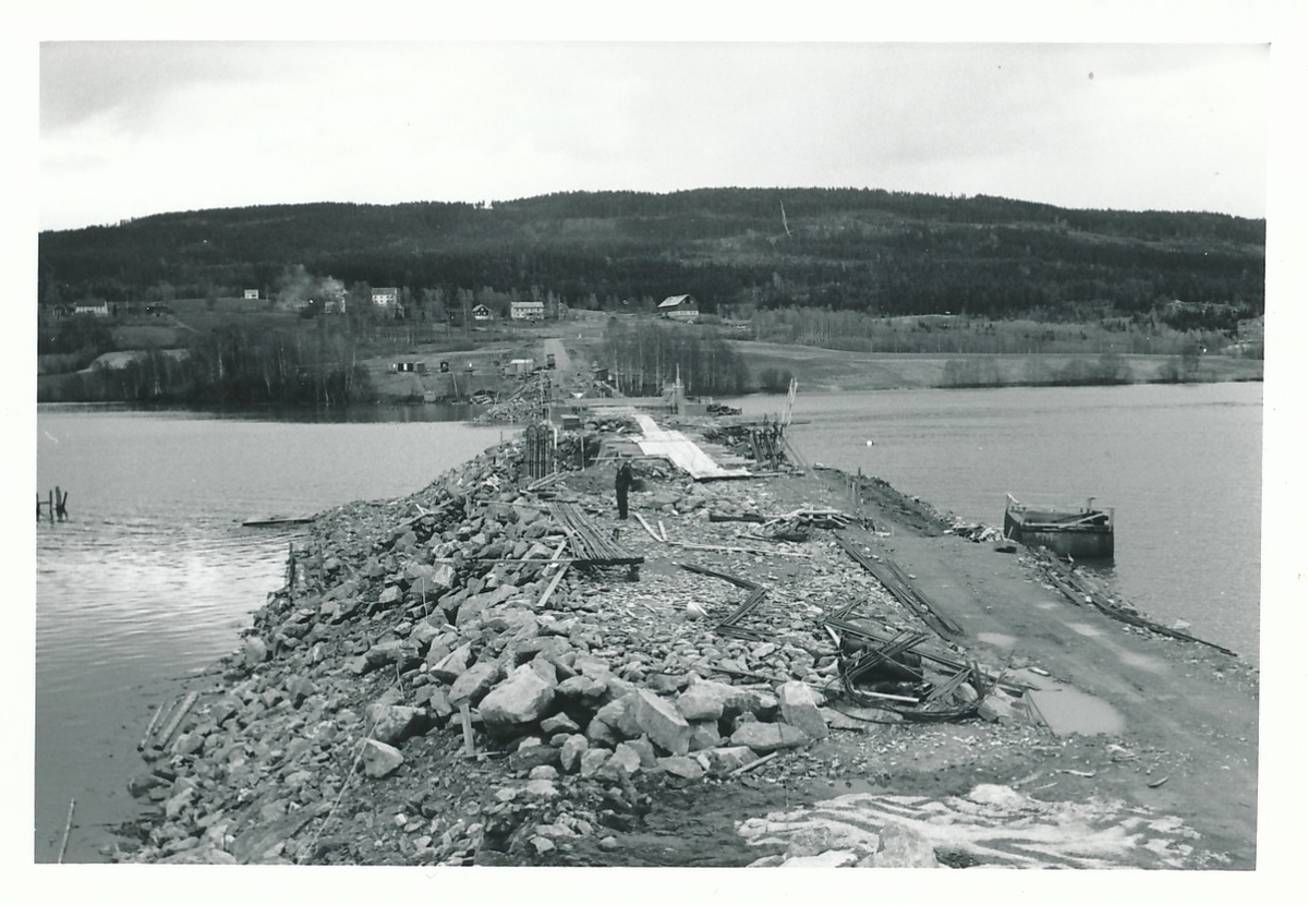Anleggsarbeid pågår for bygging av Flubergbroa over Randsfjorden. Fjorden med fyllmasser og gårder og jordbrukslandskap i bakgrunnen.