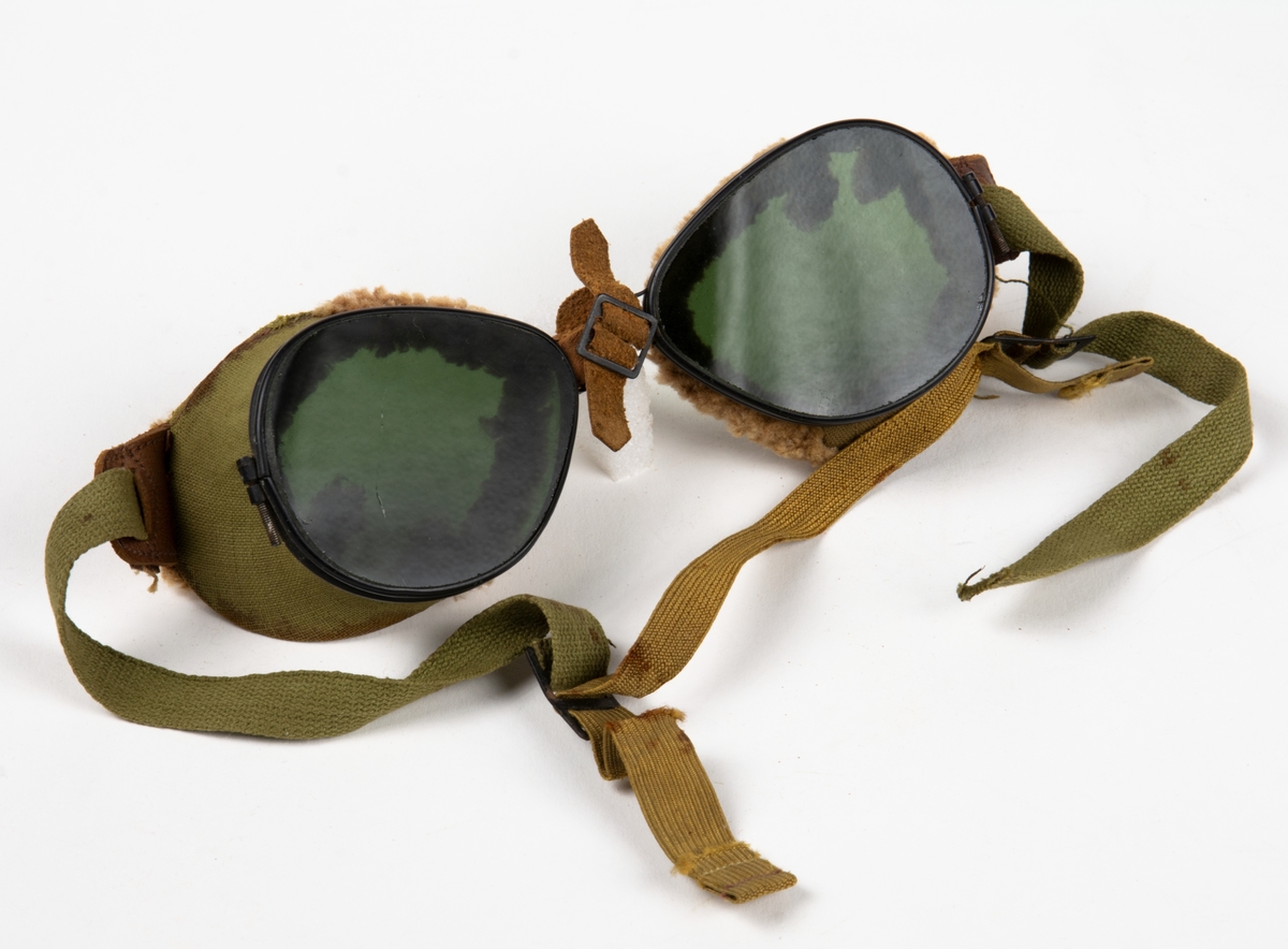Flyglasögon, med mörka glas. Glasögonens sidor är klädda med ett grovt bomullstyg i grön färg och kantade med ljusbrunt pälsfoder.
