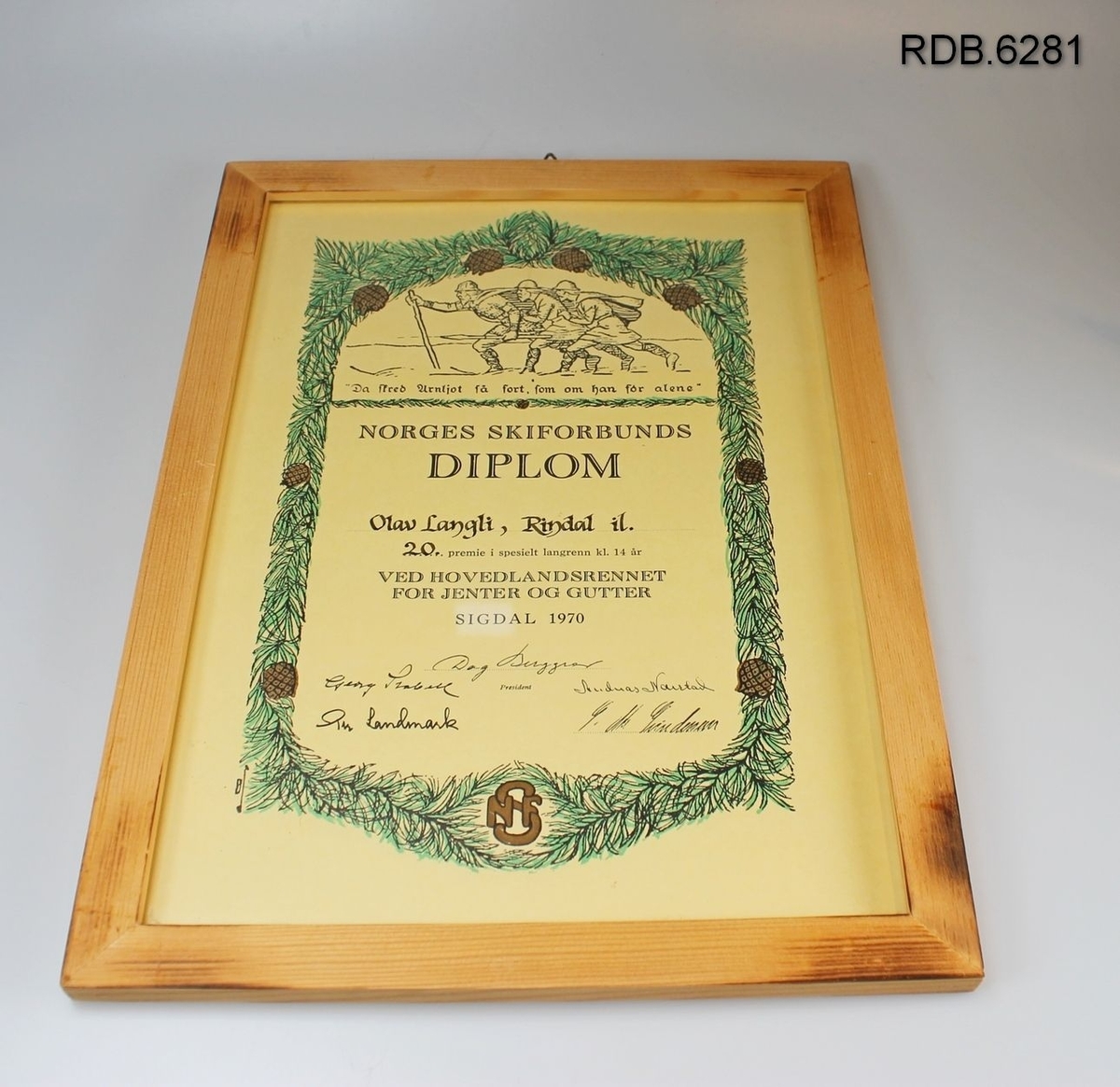 Diplom i glass og trekvit ramme. Diplomen er dekorert med en grønn krans av furubar med brune kongler og tre birkebeinere på ski øverst.
