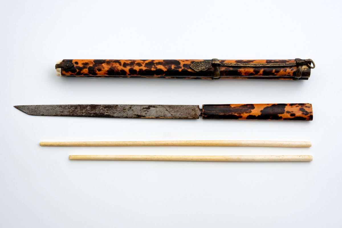Kinesisk spisesett bestående av en slire i bambus dekket med skilpaddeskjell -finér med beinbunndeksel. 
I sliren en lang kniv med skaft i samme materiale som sliren og metallblad, og et sett (2 stk) spisepinner i bein.
