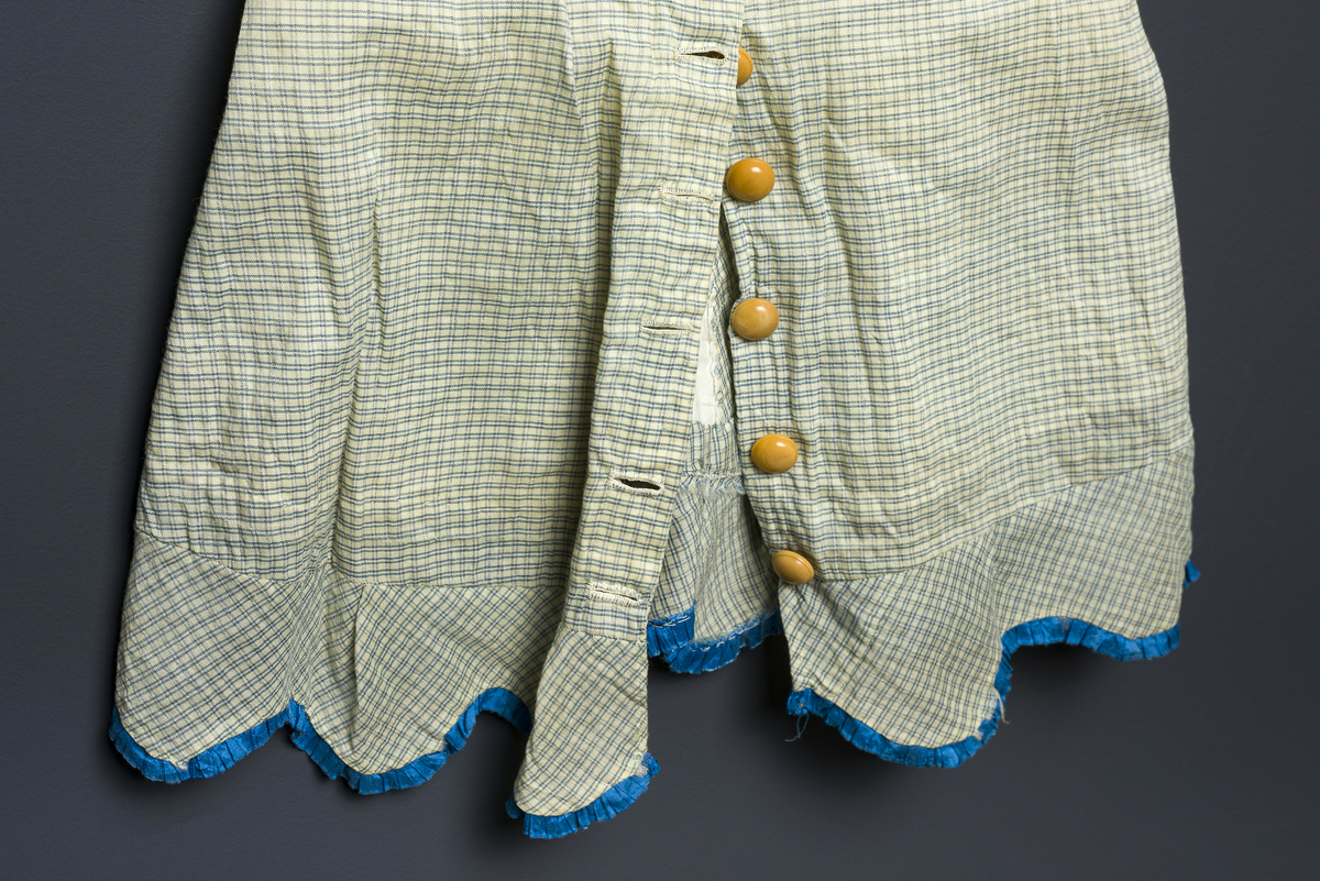 Beige og blå smårutete barnekjole med 10 brune knapper i knappestolpe bak. Dekorbånd sydd i rysjer i kantene i blått. 
Har tilhørt Sara Fabricius.
