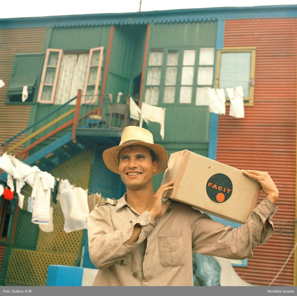 Facit-Argentina. En man med en låda Facitprodukter på axeln.I bakgrunden bostadshus där tvätten  hänger och torkar på tvättlinor.