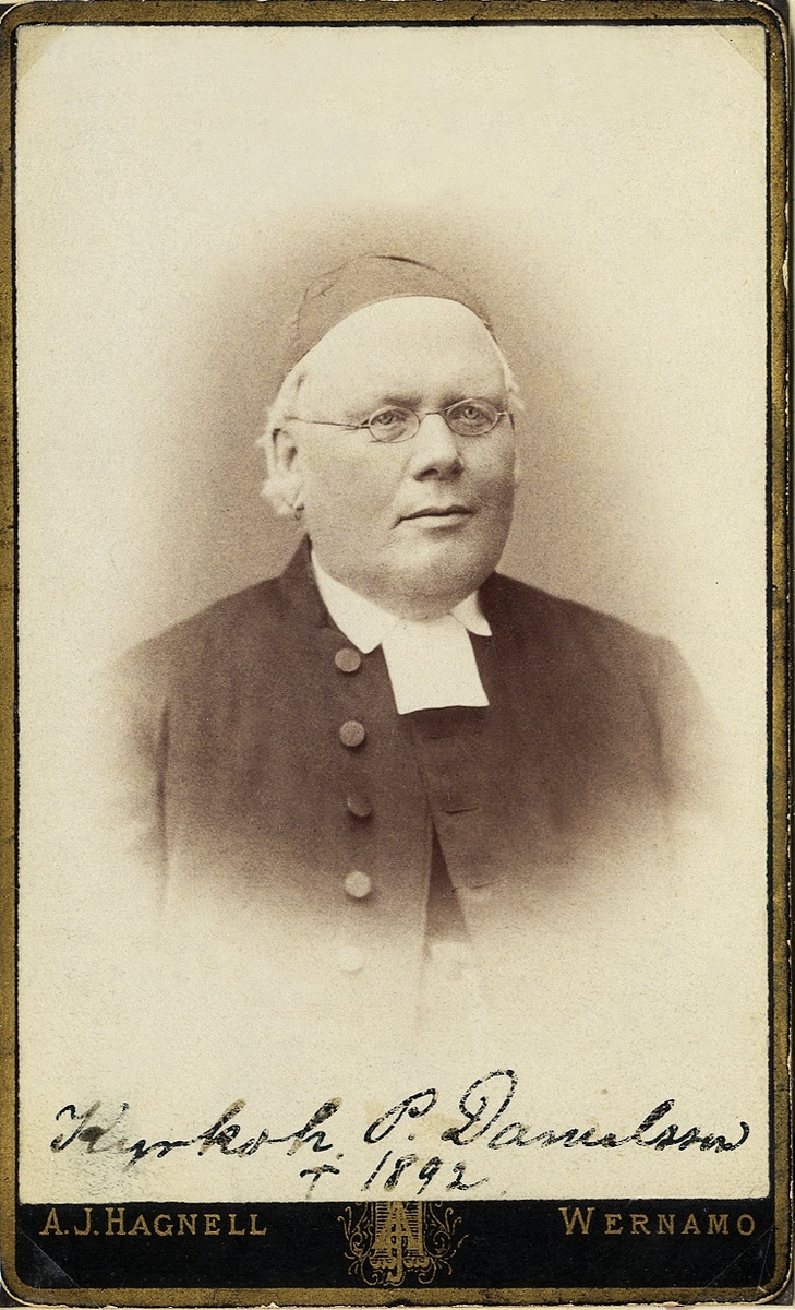 Porträttfoto av en medelålders man med glasögon, klädd i prästdräkt och kalott. 
Bröstbild, halvprofil. Ateljéfoto.