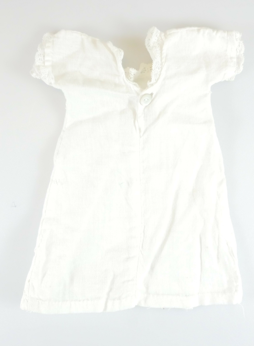 Liten hvit kjole i bomull, antageligvis brukt som dukkeklær. Korte ermer og rund hals med påsydde blondekanter. På baksiden er det en hvit knapp. 