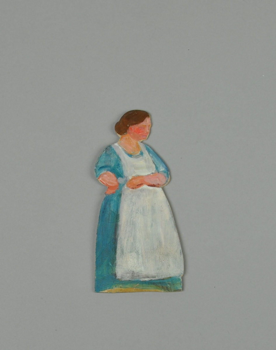 Figur av papp. Figuren forestiller dame i sideprofil, med blå kjole og hvitt forkle.