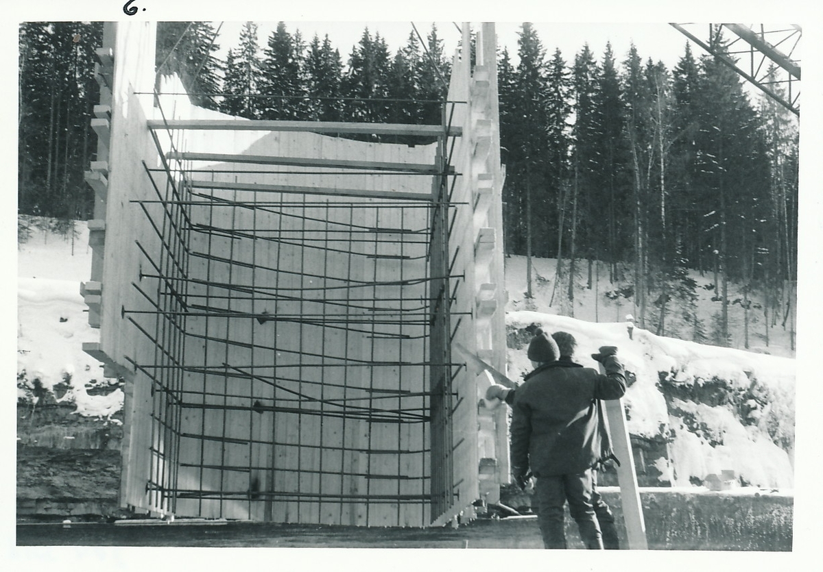 To menn står ved siden av en trekonstruksjon med tre vegger og armeringsjern inni. I bakgrunnen er Randselva og skog.