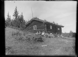 Mennesker sittende foran laftet hytte med torvtak på fjellet